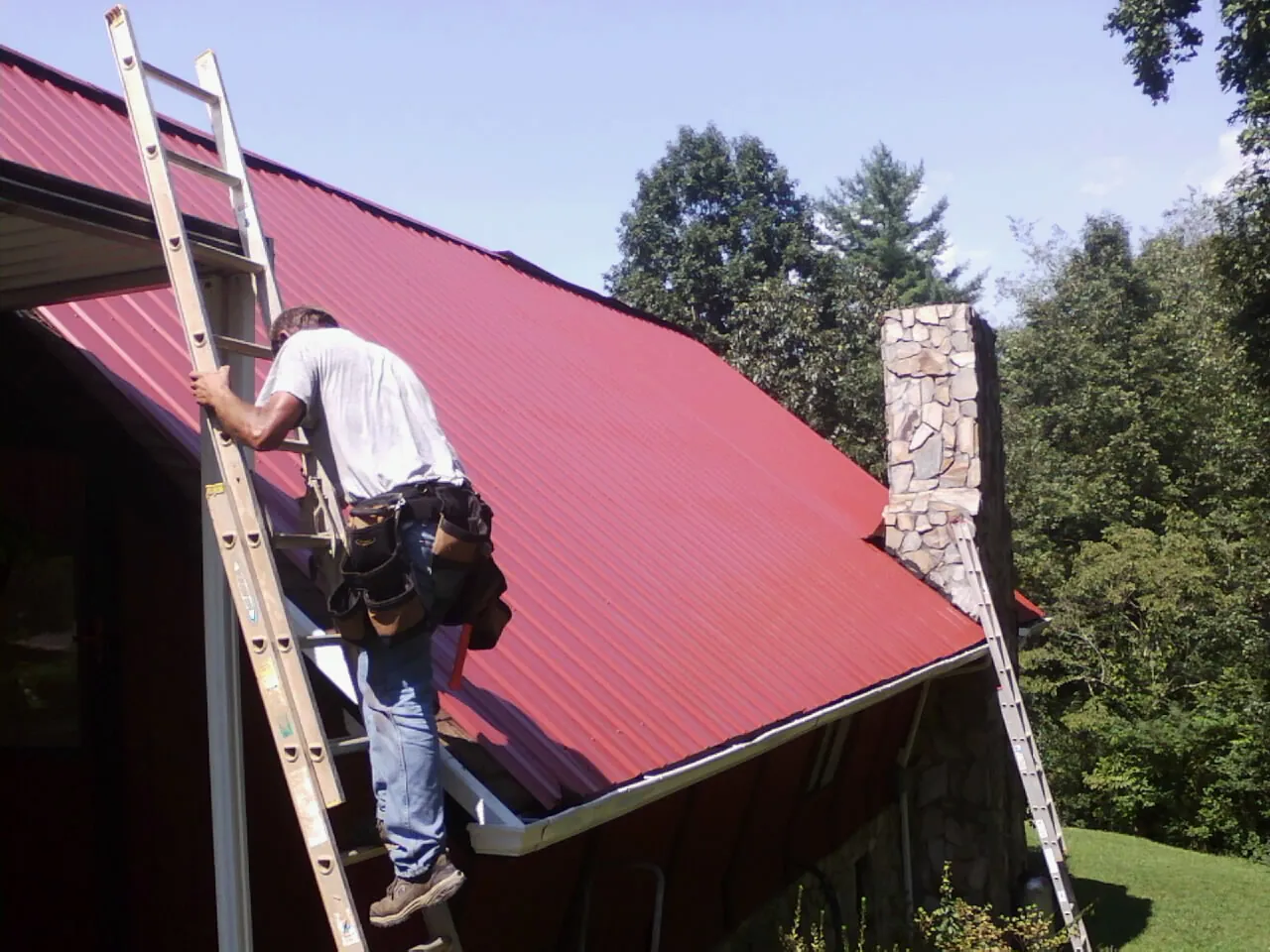 Roofer on a ladder beside a red metal roof and stone chimney — Soffit & Fascia Repair by Roofing Brothers in Maitland